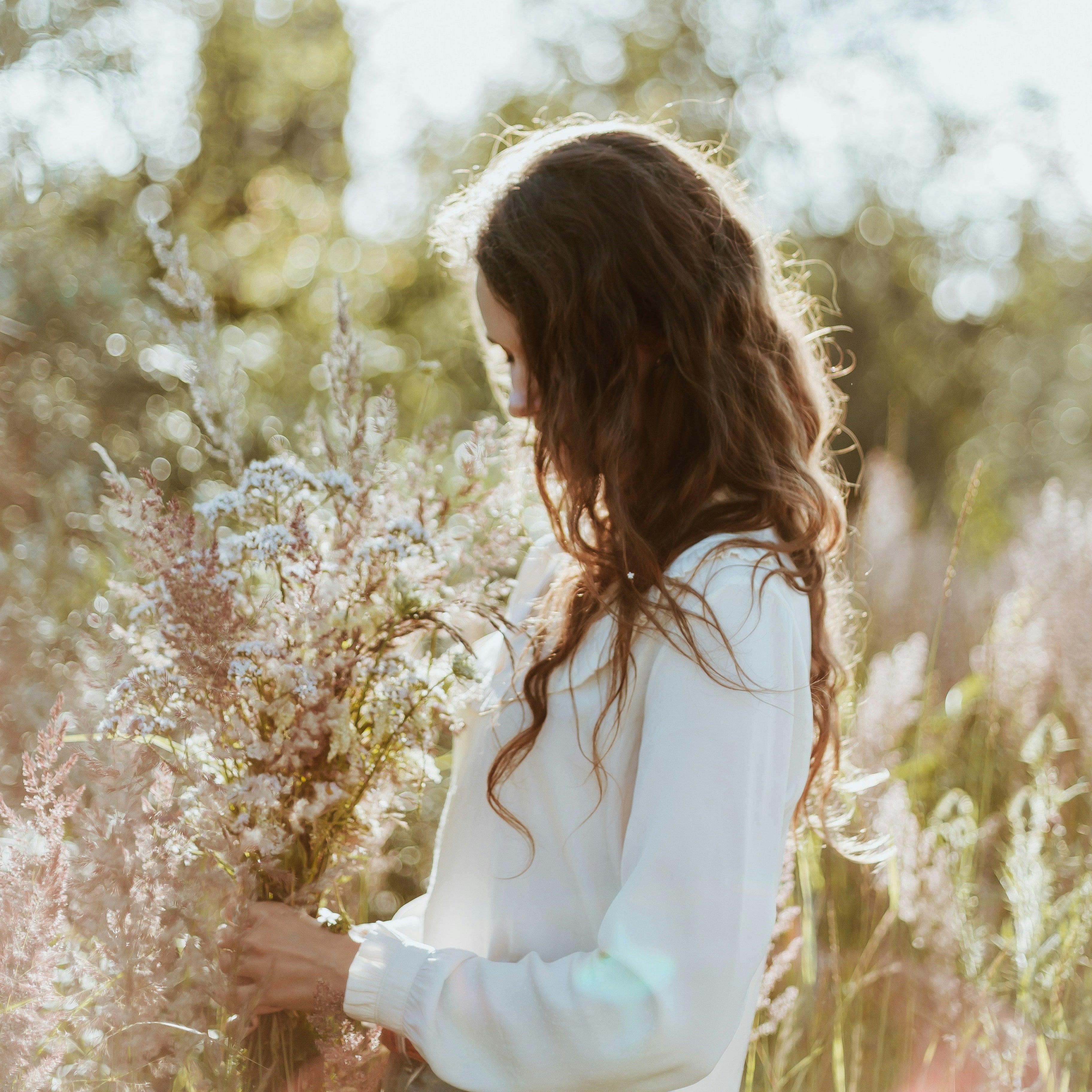 Frau in weißem Kleid hält zarten Blumenstrauß im Sonnenlicht, symbolisiert BLÜTENWEICH Duftmelts mit Kirschblüte und Reismilch Duft.