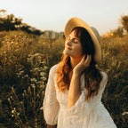 Frau mit Hut genießt Sonnenstrahlen in Blumenwiese mit Vanille-Neroli-Jasmin-Duft, Abendstimmung