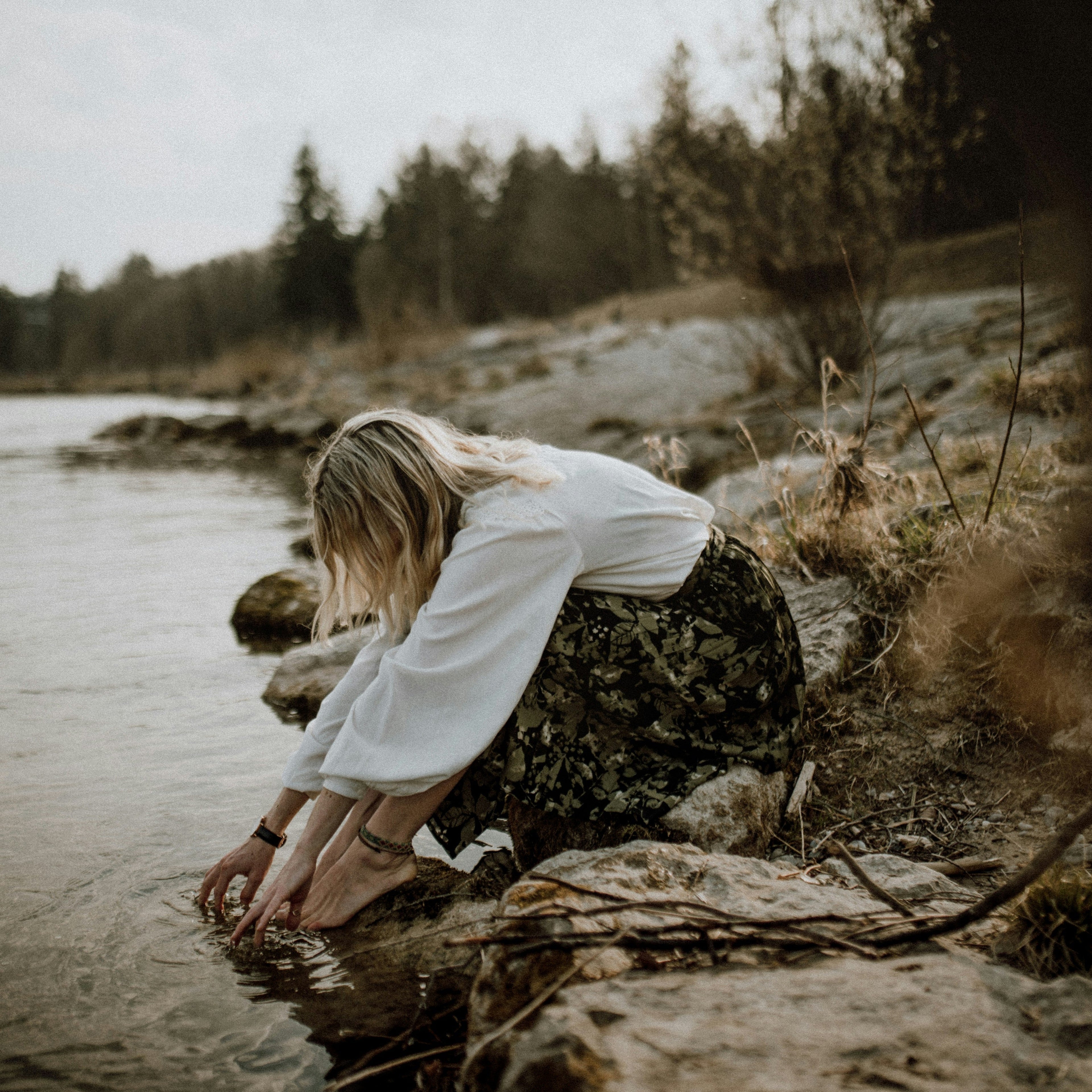 Frau am Seeufer berührt sanft das Wasser, umgeben von natürlicher Abendstimmung und ruhiger Atmosphäre, inspiriert von Duftnoten Vanille, Neroli, Bergamotte und Jasmin, Fokus auf ABENDHAUCH.
